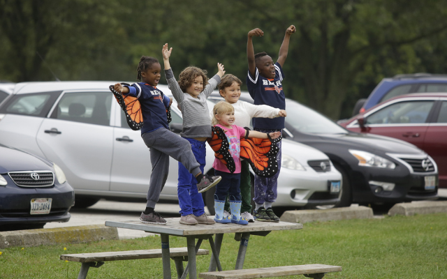 Five children stand on a picnic table with their arms outstretched as they rejoice at the Awe of Nature Festival. © Joshua Lott