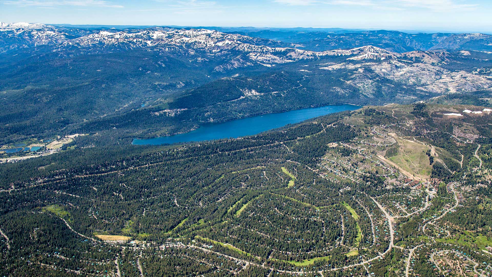 Aerial view of a narrow lake backed by snowy mountains in the distance and the winding streets of a residential subdivision built in a forest in the foreground, under clear sky with wispy clouds.