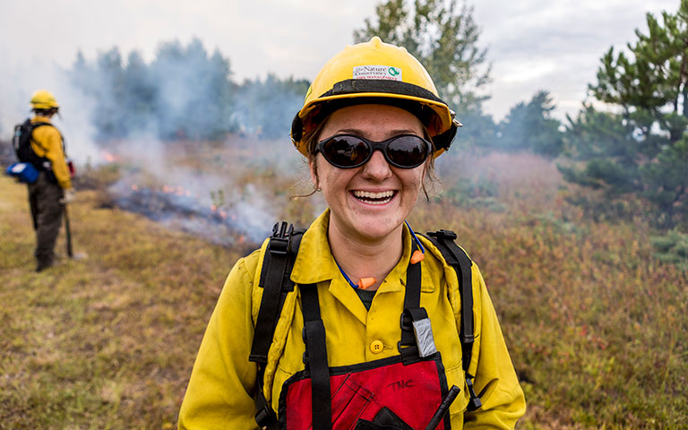 A conservation worker wearing a hard hat and sunglasses is pictured smiling in the foreground while another worker conducts a prescribed burn in a meadow in the background.