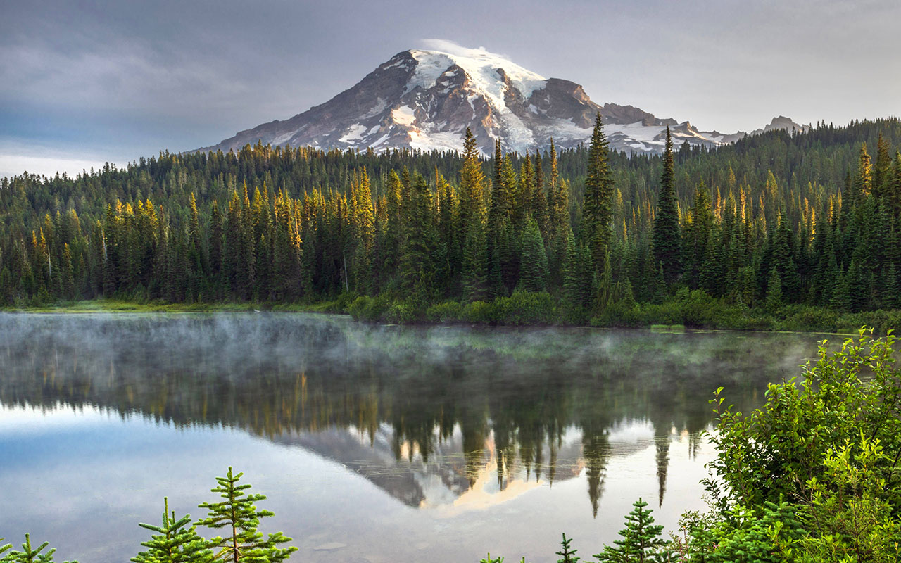 A towering, snowy and glaciated volcanic peak rises above dense evergreen forest and a small lake that reflects the mountain as mist rises from its waters.