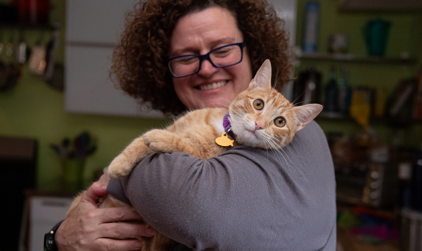 A woman in glasses holds a friendly orange cat in her kitchen.
