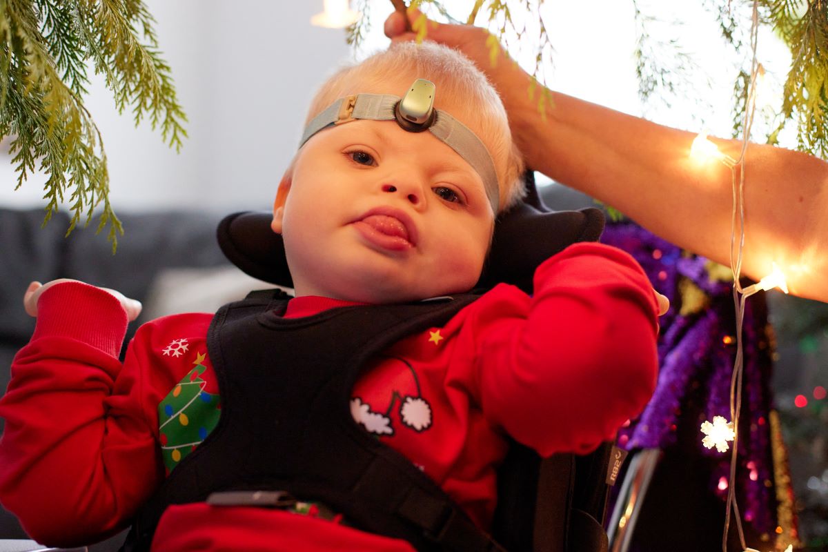 A blonde boy in a Christmas jumper sits in a supportive chair looks at the camera. Around him there are Christmas lights and you can see parts of a Christmas tree