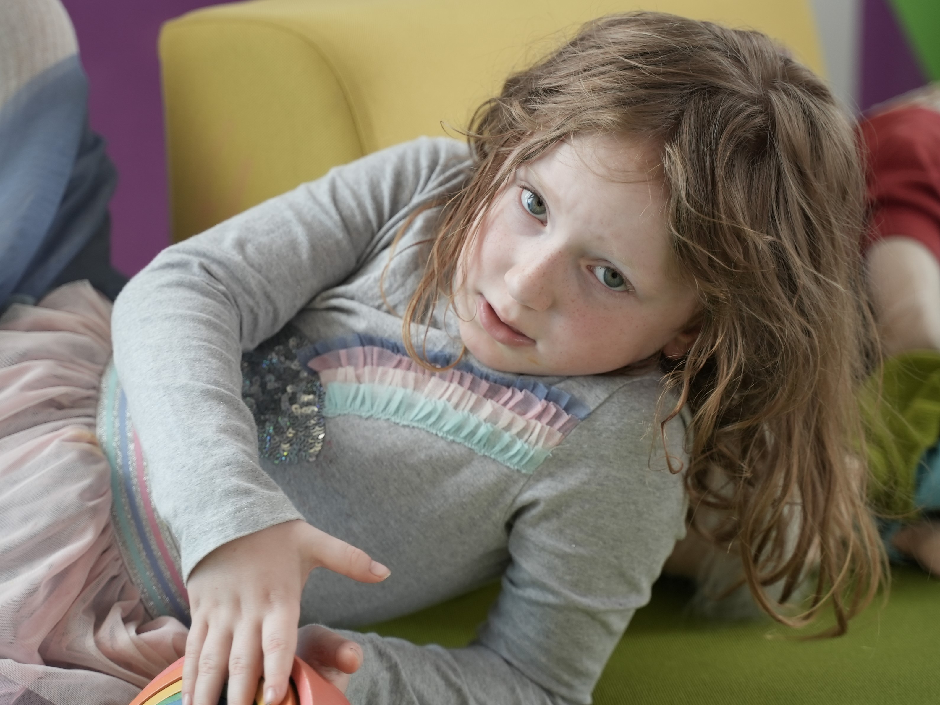 A girl with brown hair looks directly into the camera, she's wearing a grey top with a rainbow on it