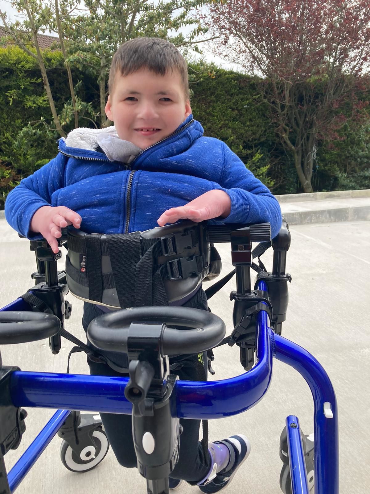 A boy wearing a blue coat uses a mobility device while smiling