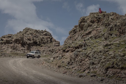 A jeep rolls along a dirt road in the African savanna, flanked to its right by the towering concrete supports of a highway