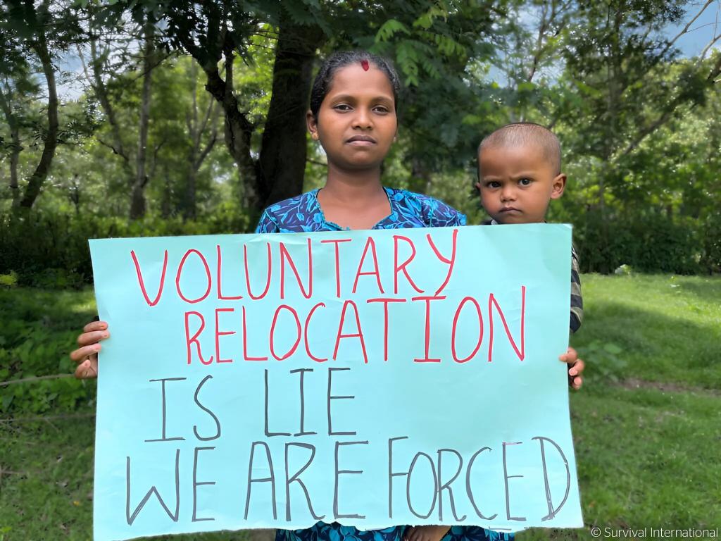 Jenu Kuruba woman and kid holding a sigh that reads "Voluntary relocation is lie. We are forced"