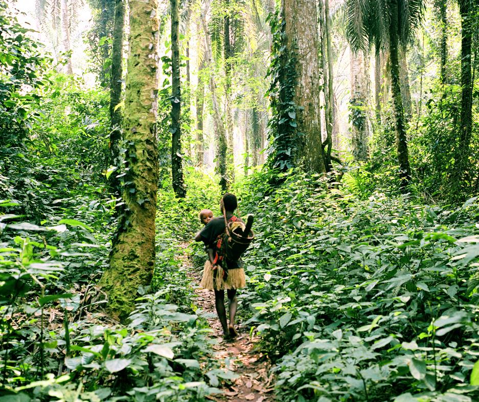 A Baka woman and child walk through lush forest.

