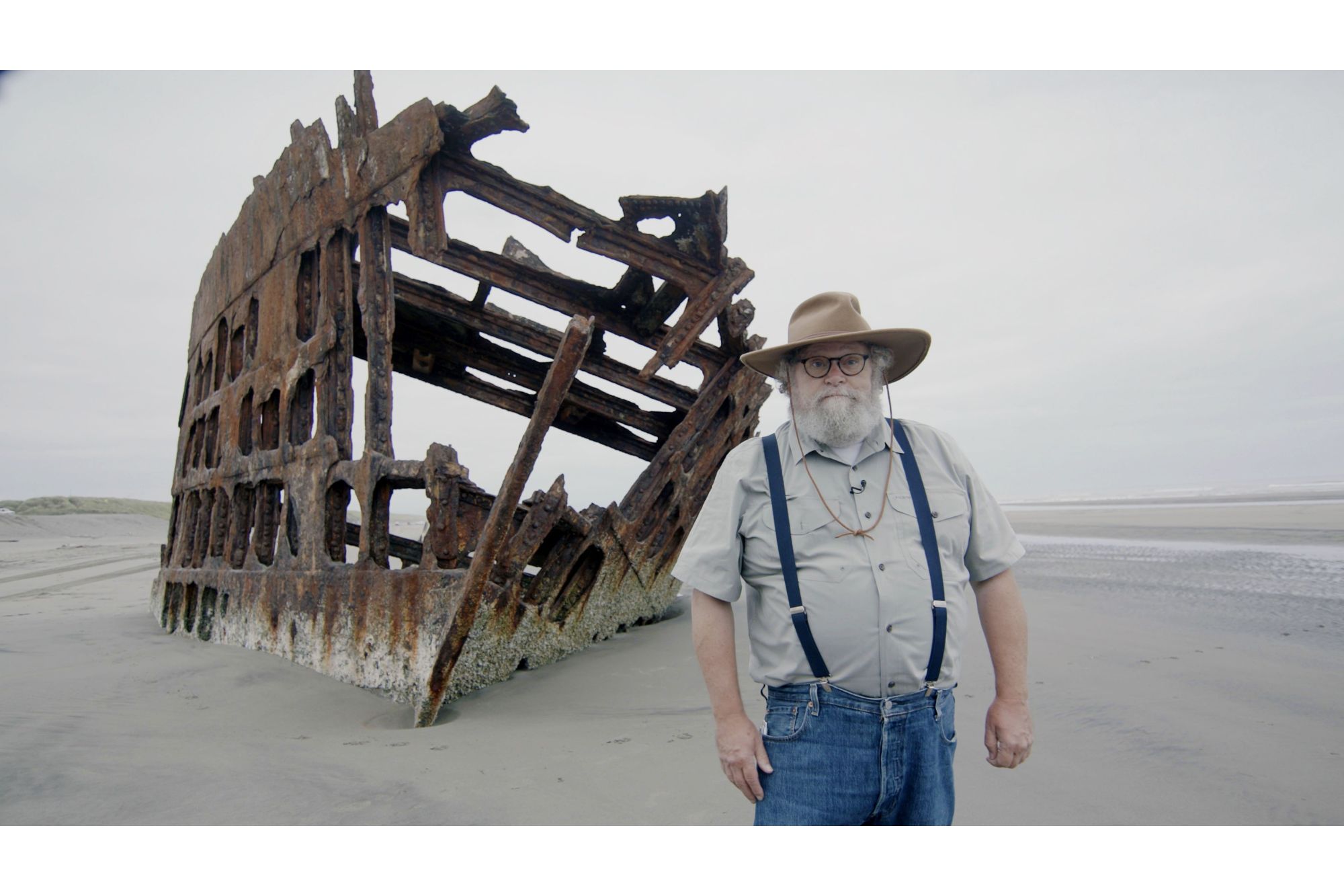 Knute Berger stands in front of a shipwreck on a beach.