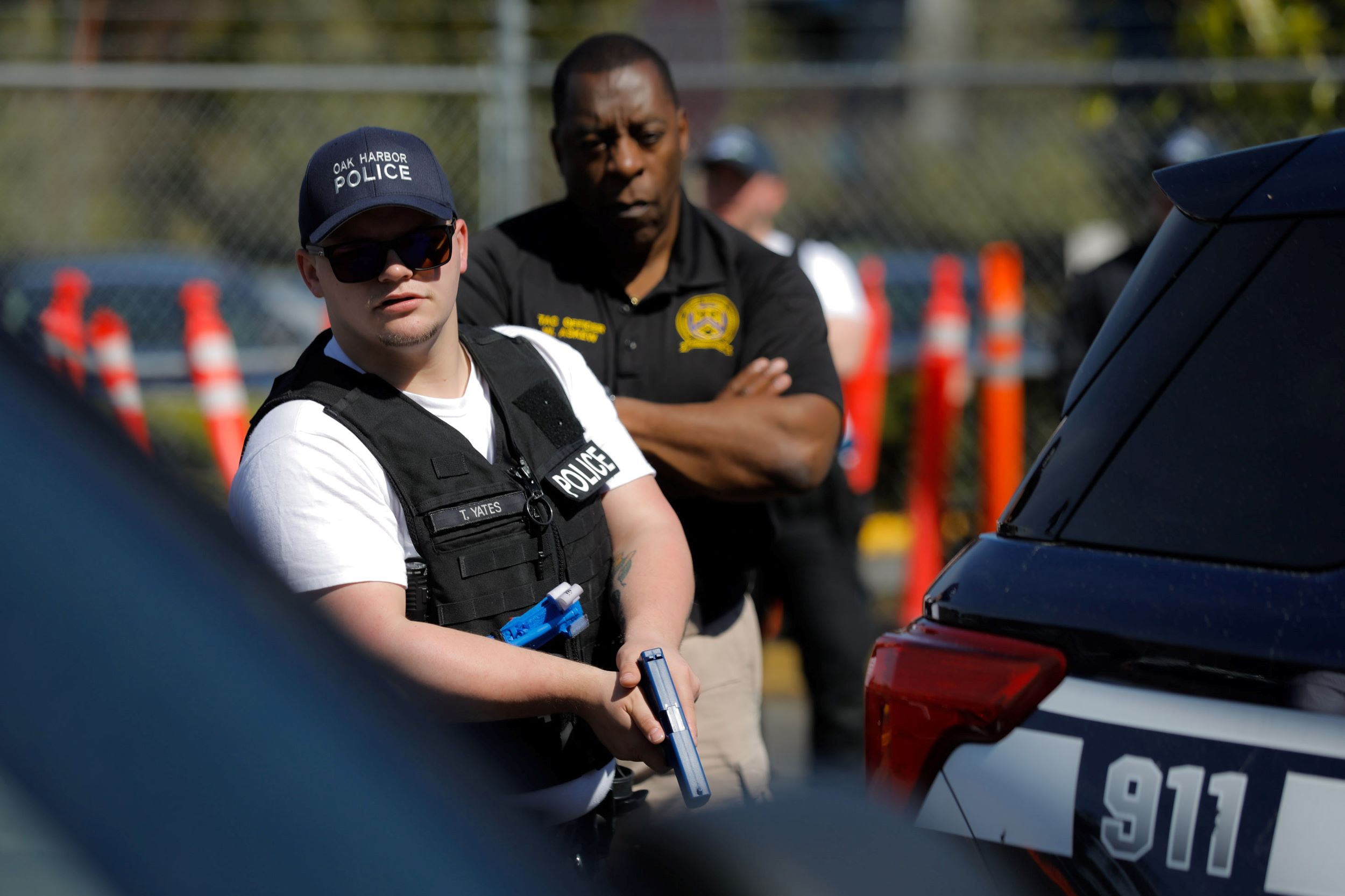 A police officer holds a gun by a cop car while another stands behind him with his arms crossed