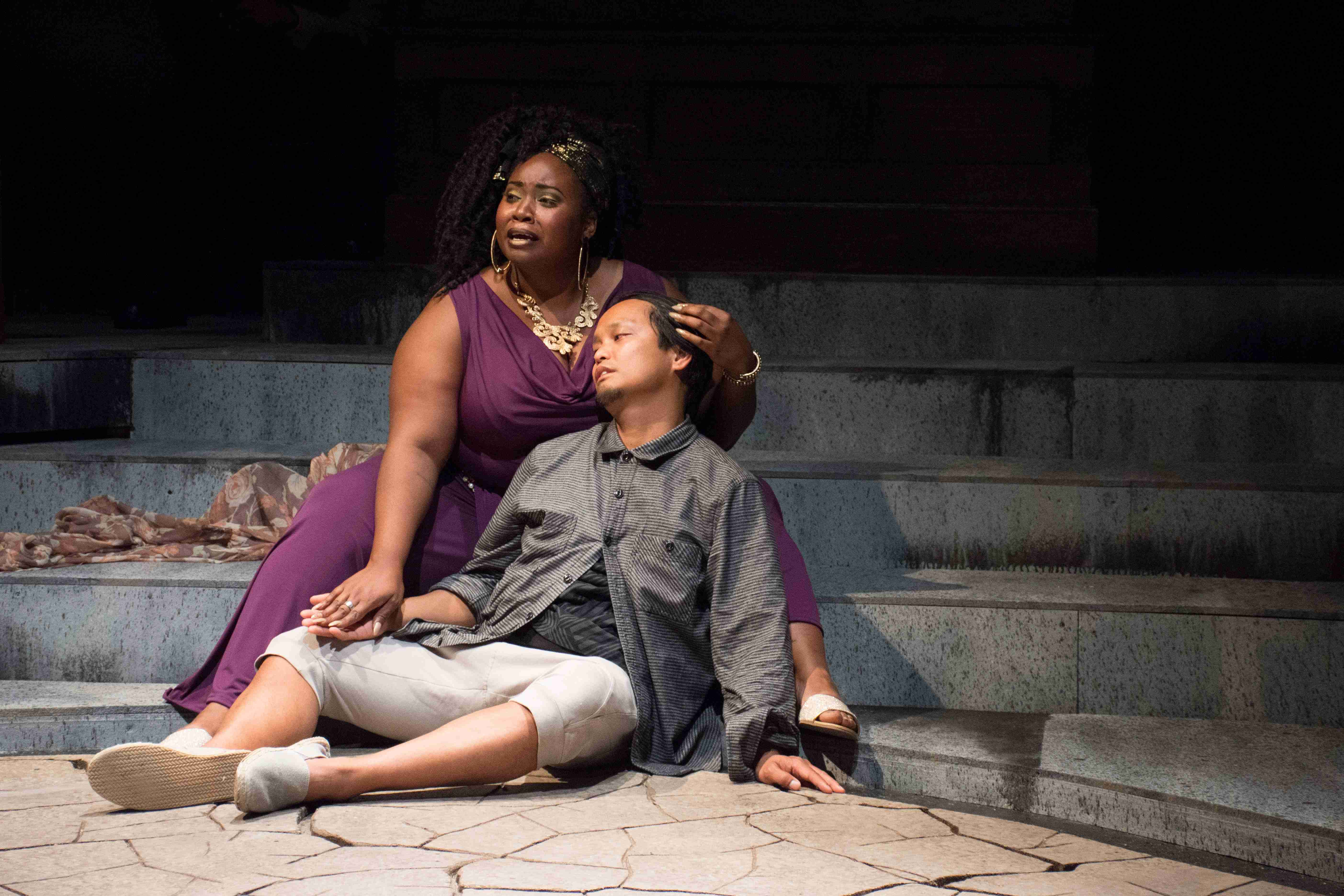 A woman and a man sit on stage during a performance, as he leans back into her lap.