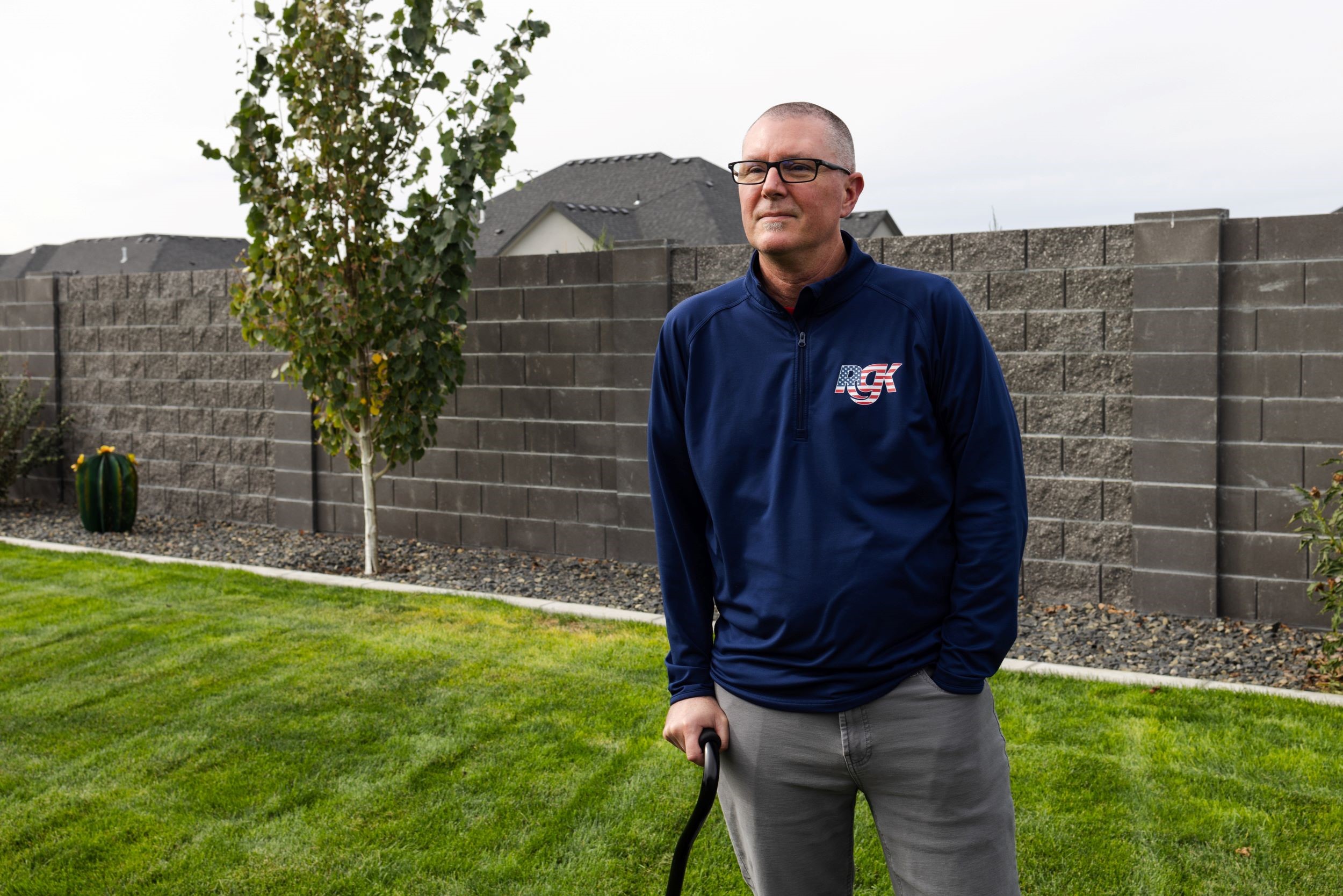 A person leaning on a cane stands on a grassy lawn in front of a gray brick wall.