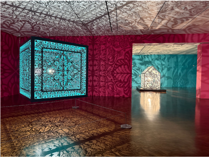 Two pink and turquoise walled gallery rooms with intricately carved installations that case shadows all over the walls.