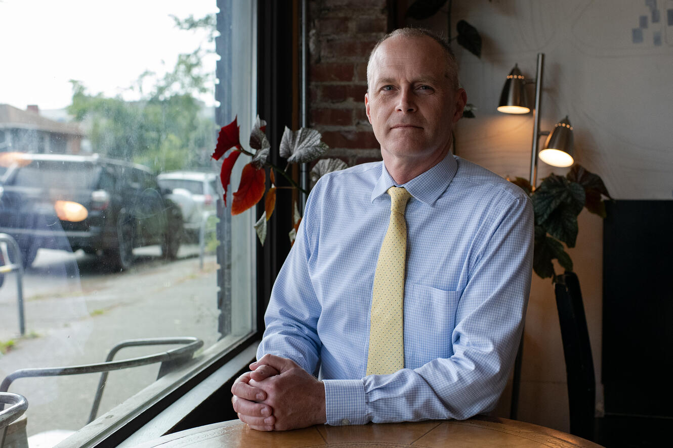 A man in a blue shirt and tie sits at a desk with his hands intertwined on the desk