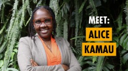 Photograph of a smiling woman standing with arms crossed in front of lush green foliage, wearing glasses, an orange blouse, and a grey blazer. Text on right side reads 
