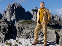 A photo of Chris Packham standing ontop of a snowy mountain, wearing a yellow outfit, with blue sky in the background.