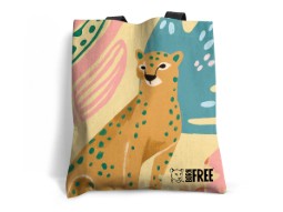 Photograph of a colourful tote bag featuring an illustrated cheetah with blue spots against abstract pastel shapes in pink, green, and blue. Bag includes black handles and a small 