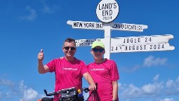 A photo of two men standing in matching pink T-shirts at the Land's End signpost, with blue sky in the background