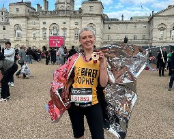 A woman runner smiling, wearing a Born Free vest holding her Marathon medal, wrapped in a foil blanket