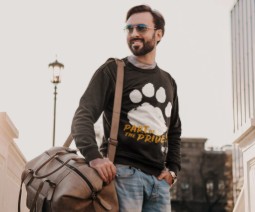 A man wearing sunglasses with a larg bag over his shoulder, standing in a residential street. His black sweatshirt has a large paw print logo and features the words:'Part of the Pride'