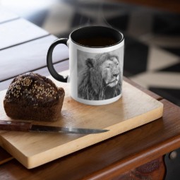 A mug featuring a black and white image of a male lion. The mug is on a coffee table with a piece of cake and a knife.