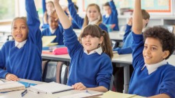 Photograph of a classroom with students wearing blue uniforms raising their hands, indicating active participation. Desks have notebooks and pencils, suggesting a learning environment focused on engagement and interaction.