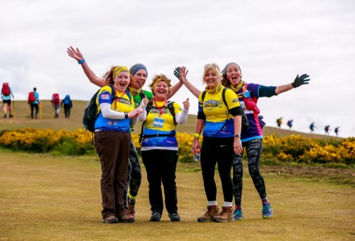 A group of women wearing brightly coloured sports clothing, standing together in a field, with walkers in the background. The women are joyfully stretching out their arms in celebration!