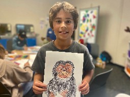 A photo of a young boy holding up his drawing of a chimpanzee, in a classroom