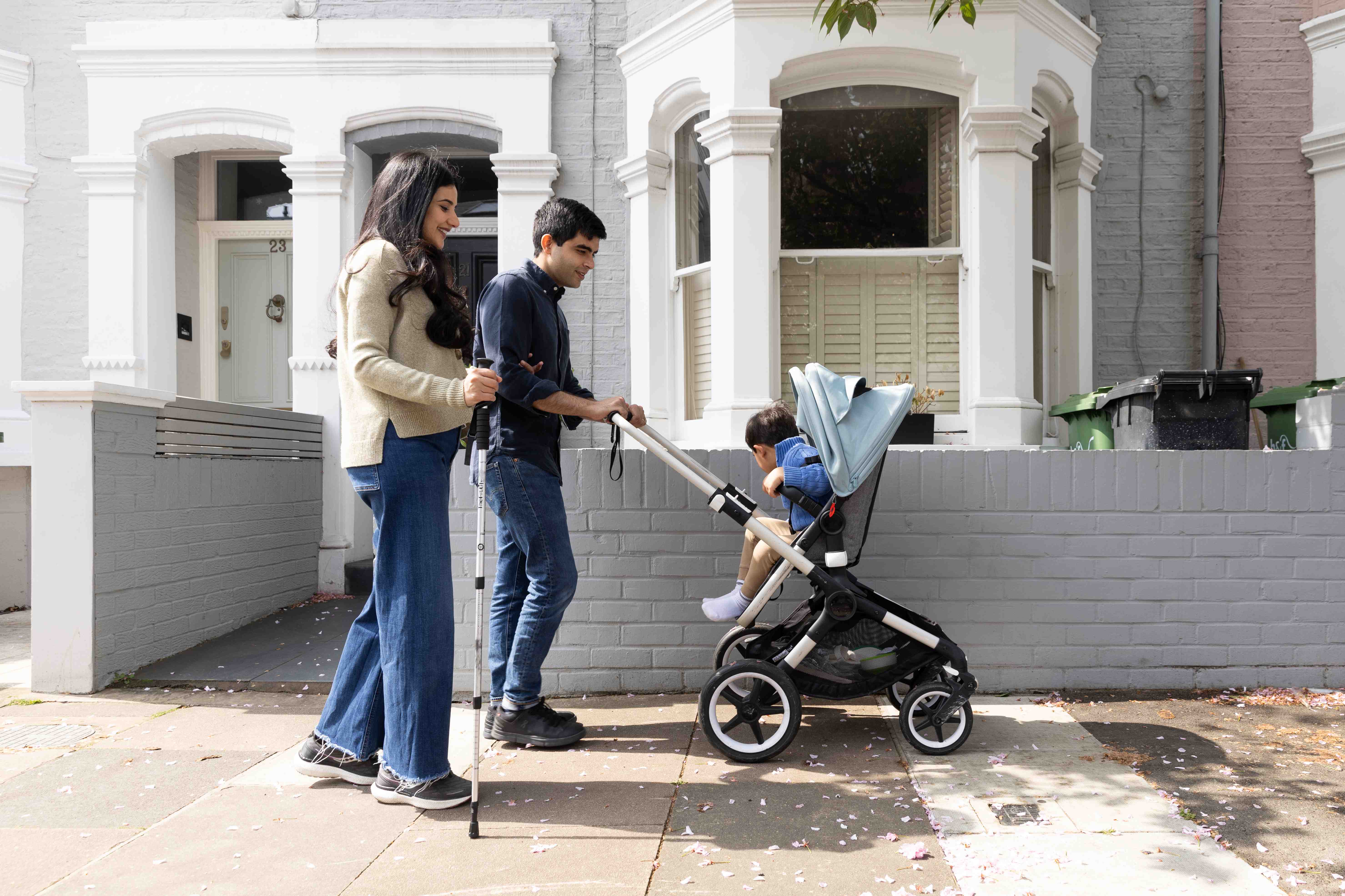 A woman holding a walking stick walking on a pavement with a man who is pushing a pram. There is a small child in the pram.